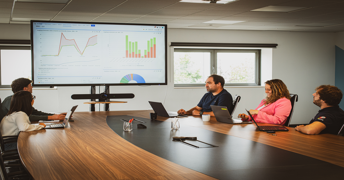 Meeting around a table, with several people exchanging in front of a large screen displaying cost-related graphs.